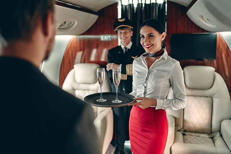 Flight attendant stands in aisle of private jet holding tray of champagne as part of in-flight private jet catering experience